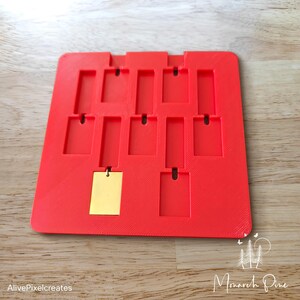May include: Red plastic mold with 12 rectangular slots for making jewelry or other crafts. A gold metal piece is in one of the slots.