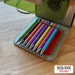 May include: A set of eight colorful pens with silver accents, arranged in a row in a metal case. The pens are green, brown, orange, purple, pink, yellow, light pink, and blue.
