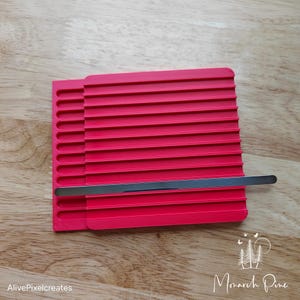 May include: A red plastic tool with a metal bar for creating parallel lines. The tool has multiple slots for creating different widths of lines.