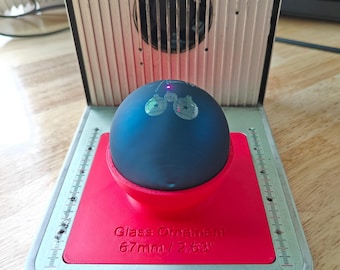May include: A blue glass ornament with a white design of two Christmas bells. The ornament is sitting on a red platform with the text "Glass Ornament 67mm/2.63" printed on it.