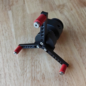 May include: Black tripod with three adjustable legs, each with a red rubber foot. The tripod has a black circular base with a small black arm extending from the top.