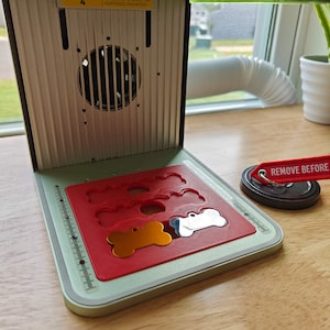 Puede incluir: Una máquina verde y plateada con una bandeja roja que contiene varias etiquetas de metal en forma de hueso. La bandeja tiene una regla en el lateral. Una cinta roja con el texto "REMOVE BEFORE" es visible en el fondo.