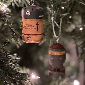 May include: Two Christmas ornaments hanging on a green Christmas tree. The first ornament is a brown and gray cylinder with red and white text that reads "WARNING!  THIS ITEM MAY EXPLODE IF HEATED COLD OR HOT". The second ornament is a gray and brown rocket with a red top.