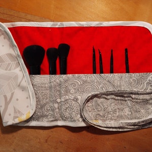 Puede incluir: Un rollo de brochas de maquillaje de tela blanca y gris con estampado paisley y forro rojo. El rollo está enrollado y atado con una cinta blanca. Hay cinco brochas de maquillaje negras dentro del rollo.