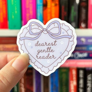May include: A heart-shaped sticker with a lavender bow and the words "dearest gentle reader" in cursive. The sticker has a scalloped edge and small star and flower accents. The background features a shelf of colourful books.