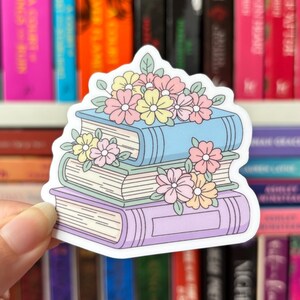 May include: A sticker depicting a stack of three books in pastel shades: light blue, green, and lavender. The books are decorated with pink and yellow flowers and green leaves, creating a charming illustration.