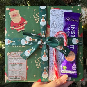 May include: A festive Christmas gift box wrapped in green paper with Santa Claus and snowflake patterns. It's tied with a green ribbon with Santa Claus designs. Inside are a Cadbury hot chocolate sachet, a candy cane, and a coloring card.