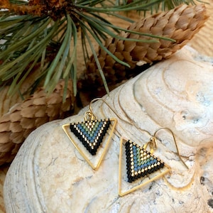 May include: A pair of gold-tone triangle earrings with a beaded design in black, blue, and green. The earrings are displayed on a textured, off-white surface, with pine needles and a pine cone in the background.