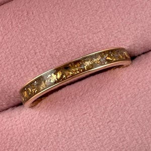 May include: A gold-colored ring with a band filled with gold and amber-colored flecks. The ring is set inside a pink velvet ring box, with a matching pink background.