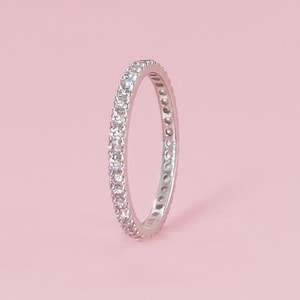 May include: A silver-toned eternity band ring featuring a continuous row of small, clear, round-cut stones set along the entire outer circumference of the metal band. The ring is positioned upright against a soft, light pink background.