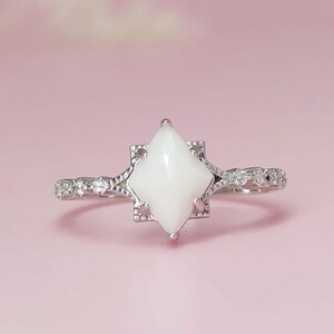 May include: A silver ring featuring a white, diamond-shaped central stone. The band is adorned with small, clear gemstones. The ring is set against a soft pink backdrop.