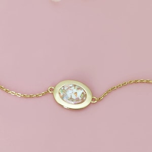 Könnte beinhalten: Ein goldfarbenes Armband mit einem ovalen Anhänger, der eine Mischung aus weißen und klaren Steinen enthält. Das Armband liegt auf einem rosa Hintergrund mit einem Zweig getrockneter Blumen.