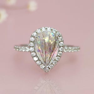 May include: A silver ring with a pear-shaped, clear gemstone, surrounded by a halo of small, clear stones. The gemstone has internal, colorful inclusions. The ring band is also set with small, clear stones. The background is a soft pink.