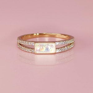 Op de afbeelding: Een goudkleurige ring met een rechthoekige opaal middensteen. De band splitst zich in twee rijen, elk bezet met kleine, heldere edelstenen. De ring staat tegen een zachte roze achtergrond, wat het elegante ontwerp benadrukt.