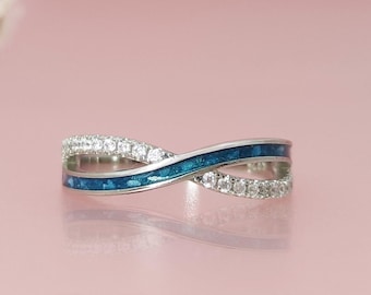 May include: A silver-toned ring with an infinity design. The ring features a band of sparkling clear stones and a band of blue enamel. The ring is displayed on a pink surface.