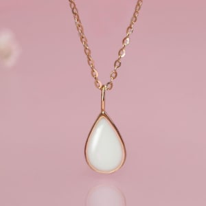 May include: A gold-colored necklace with a teardrop-shaped pendant. The pendant has a white, pearlescent center and a gold-colored frame. The necklace hangs against a soft pink background.