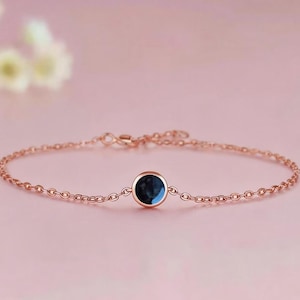 May include: Rose gold bracelet with a delicate chain and a single, round, dark blue gemstone. The bracelet is displayed on a soft pink background, with a blurred floral element in the upper left corner. The bracelet's chain is made of small, interconnected links.