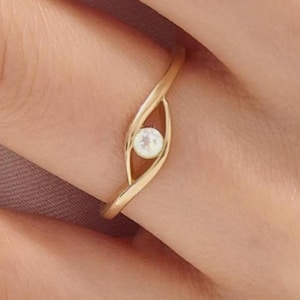 May include: A delicate gold ring featuring a small, round, white gemstone set in a unique, eye-shaped design. The ring's band curves gracefully around the finger, creating an elegant and minimalist aesthetic. The ring is a piece of jewelry.