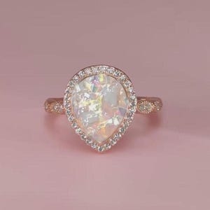 Op de afbeelding: Een roségouden ring met een peervormige, iriserende witte edelsteen in het midden. De edelsteen is omgeven door een halo van kleine, heldere stenen. De band van de ring heeft ook kleine, heldere stenen. De ring staat tegen een roze achtergrond.