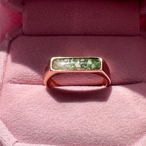 Peut inclure: Une bague chevalière en or rose avec une pierre rectangulaire verte et blanche, présentée dans un écrin en velours rose. L'écrin est ouvert, révélant la bague. Le texte "LOVE CATCHER" est visible à l'intérieur de l'écrin.