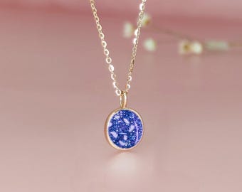 May include: A gold-colored necklace with a round pendant. The pendant features a dark blue speckled design with white accents. The necklace is displayed against a soft pink background, highlighting the jewelry's delicate features.