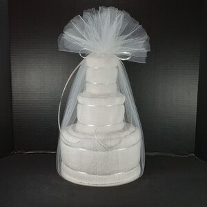May include: A white towel cake wrapped in white tulle with a white ribbon. The cake is made of four towels stacked on top of each other.