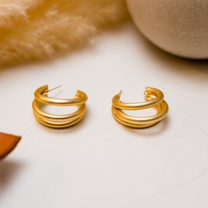 May include: A pair of gold hoop earrings with a triple layered design. The earrings are on a white surface.