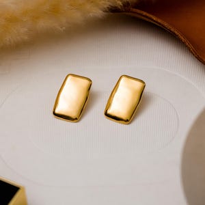 Puede incluir: Un par de pendientes rectangulares de color dorado. Los pendientes son simples y de diseño moderno.