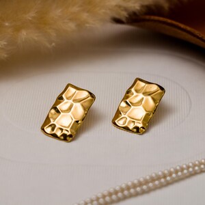 May include: A pair of gold-toned earrings with a textured, geometric design. The earrings are rectangular in shape and have a hammered finish.