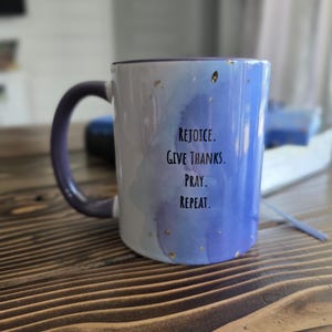 May include: A ceramic mug with a purple handle and rim. The mug features a watercolor design in shades of blue and purple, with gold flecks. The words "REJOICE. GIVE THANKS. PRAY. REPEAT." are printed on the side.