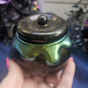 May include: A small, round, iridescent green and black cauldron-shaped container with a matching lid. The lid is topped with a small, round knob. The container has a glittery, speckled finish. The cauldron is held in a person's hand.