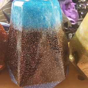 May include: A faceted, decorative resin tumbler with a gradient of colors. The top is a sparkling teal, transitioning to a brown, glitter-infused middle, and ending with a dark blue base. The tumbler is likely used for beverages.