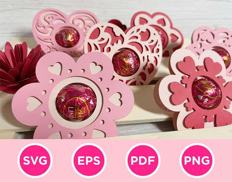 May include: Pink and red paper flower-shaped holders with heart cutouts, each holding a wrapped chocolate. The holders are arranged on a light-colored surface, with a paper flower in the background. The image includes the text "SVG", "EPS", "PDF", and "PNG".