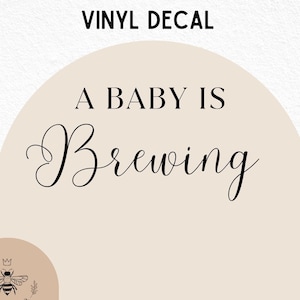 May include: A beige vinyl decal with a rounded arch shape. The decal features the text "A Baby Is Brewing" in black cursive font. A small bee logo with a crown is in the lower left corner.