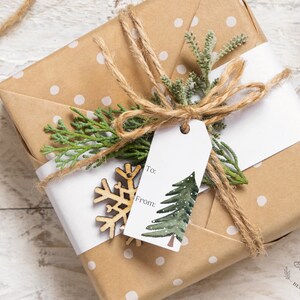 May include: A brown gift box wrapped in white polka dot paper with a white gift tag that says "To:" and "From:" and a green watercolor pine tree illustration. The box is tied with twine and decorated with a wooden snowflake and a sprig of greenery.