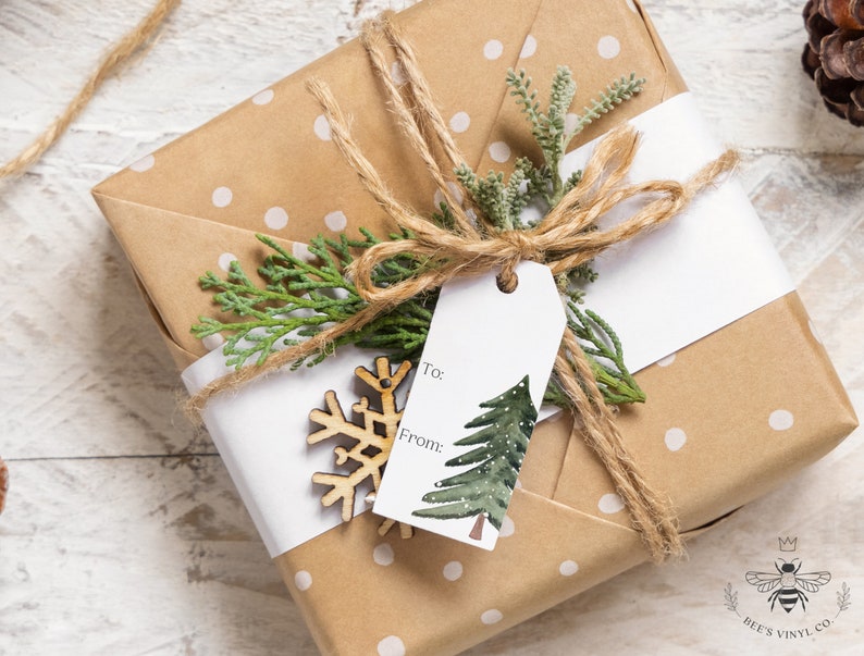 May include: A brown gift box wrapped in white polka dot paper with a white gift tag that says "To:" and "From:" and a green watercolor pine tree illustration. The box is tied with twine and decorated with a wooden snowflake and a sprig of greenery.