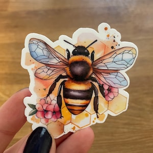 May include: A watercolor sticker featuring a bee with yellow and black stripes, translucent wings, and a honeycomb background. Pink flowers are on either side of the bee.
