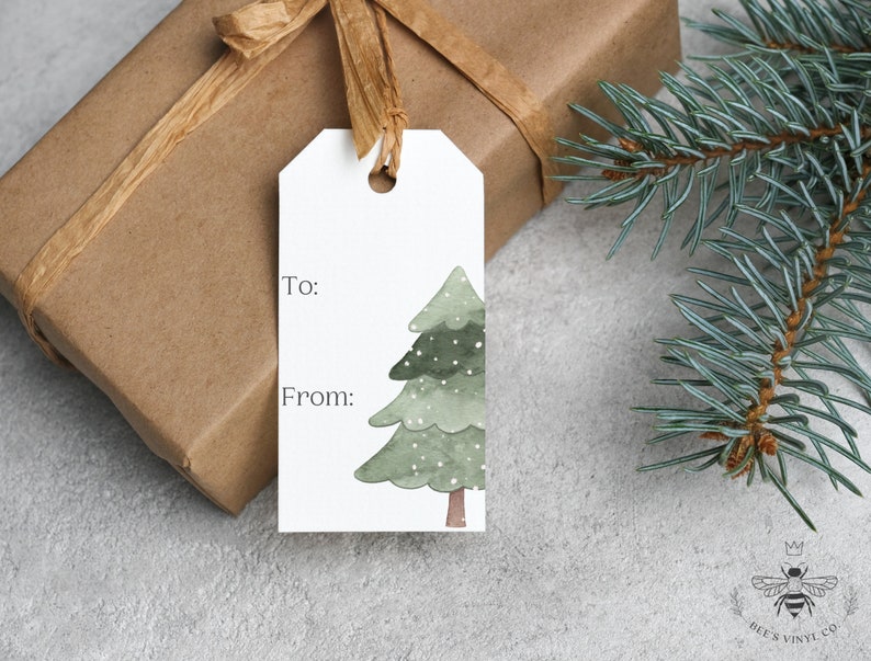 May include: A white gift tag with a watercolor illustration of a green Christmas tree with white snowflakes. The tag has the words "To:" and "From:" printed on it.
