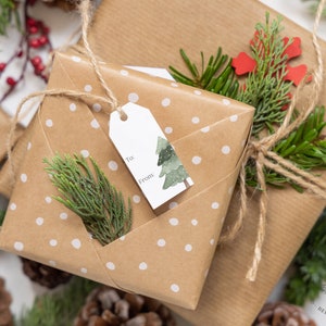 May include: A brown gift box with white polka dots wrapped with twine and a gift tag that says "To" and "From". The box is decorated with a small green pine tree and a sprig of greenery.
