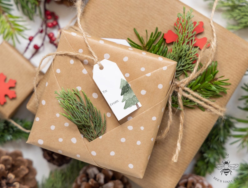 May include: A brown gift box with white polka dots wrapped with twine and a gift tag that says "To" and "From". The box is decorated with a small green pine tree and a sprig of greenery.