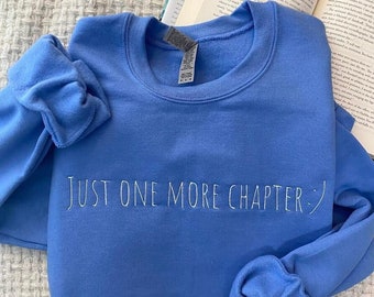 May include: A blue sweatshirt with the text "JUST ONE MORE CHAPTER" in white lettering. The sweatshirt is folded and lying on a white textured surface.