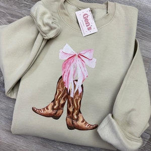 May include: A light beige sweatshirt with a graphic of brown cowboy boots tied together with a pink bow. The sweatshirt has a crew neck and long sleeves.