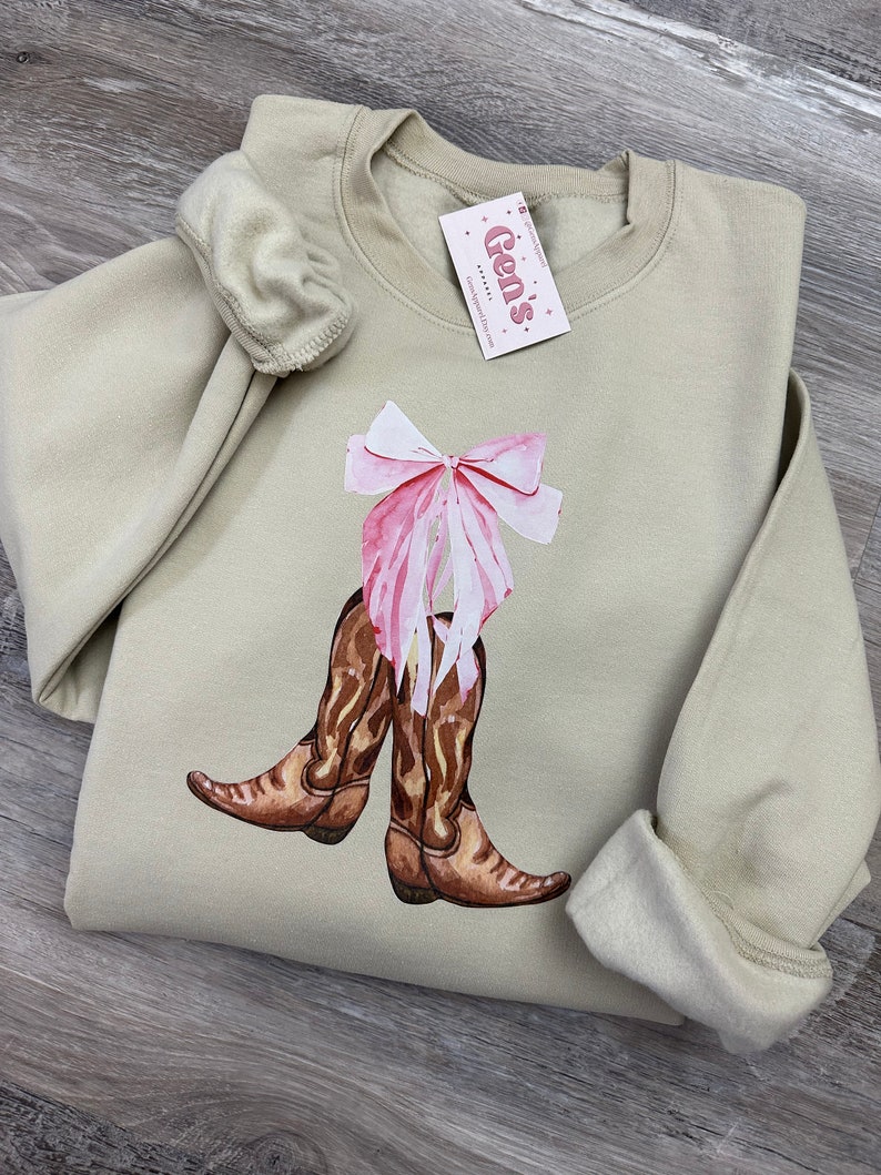 May include: A light beige sweatshirt with a graphic of brown cowboy boots tied together with a pink bow. The sweatshirt has a crew neck and long sleeves.