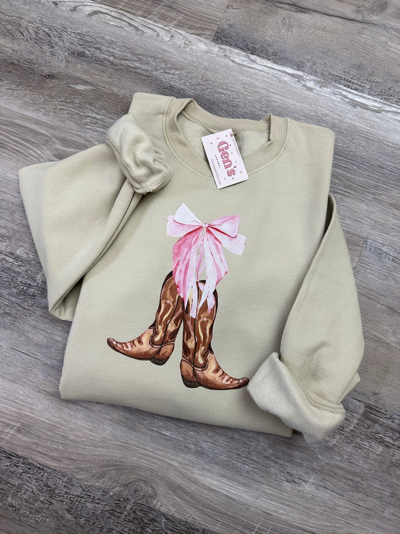 May include: A light green sweatshirt with a pink bow and brown cowboy boots printed on the front. The sweatshirt has a crew neck and long sleeves. The text "Gen's" is printed on a pink tag attached to the sweatshirt.