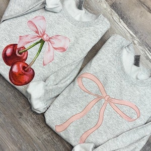 May include: Two gray sweatshirts with pink bows and cherries printed on them. The sweatshirt on the left has a cherry with a bow on top. The sweatshirt on the right has a large bow.