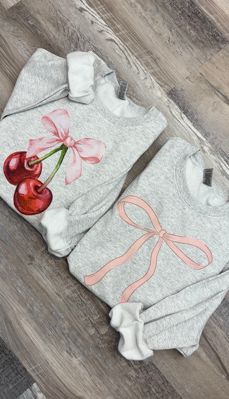 May include: Two gray sweatshirts with pink bows and cherries printed on them. The sweatshirt on the left has a cherry with a bow on top. The sweatshirt on the right has a large bow.