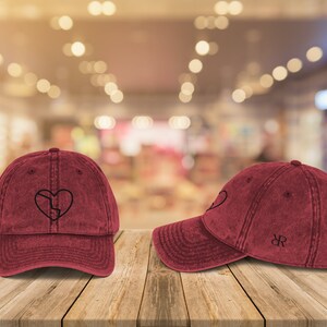 Puede incluir: Dos gorras de béisbol rojas con un aspecto desgastado. Una gorra tiene un corazón roto bordado en negro. La otra gorra tiene un logotipo "JR" bordado en negro.