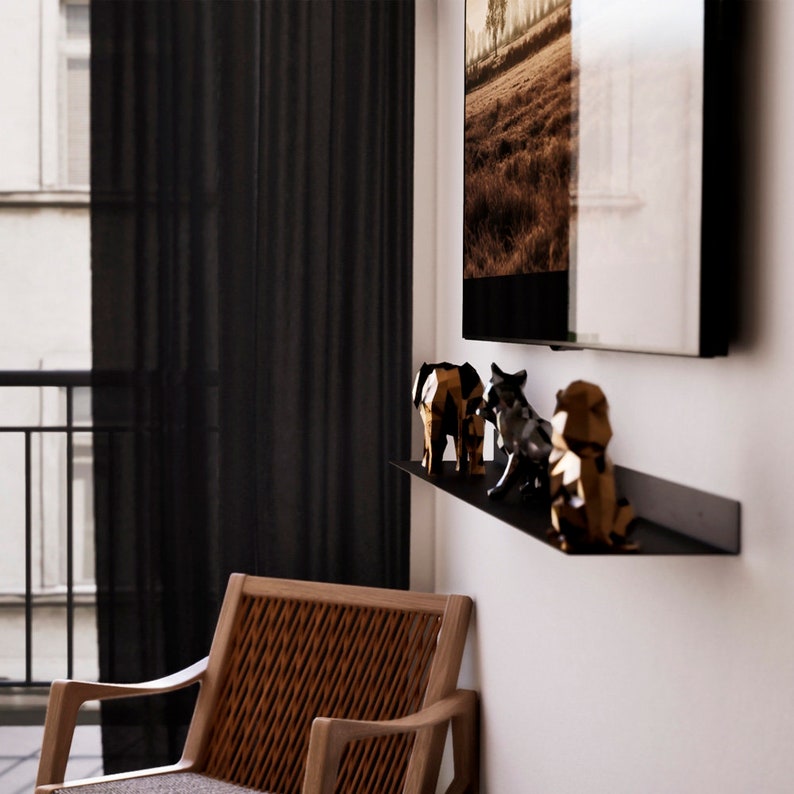 May include: A modern interior scene featuring a black wall-mounted shelf with geometric animal figurines. The figurines include an elephant, a wolf, and a lion. A framed landscape art piece hangs above the shelf, and a wooden chair is in the foreground.
