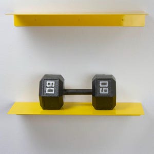 May include: A black 60-pound dumbbell rests on a bright yellow shelf, mounted on a white wall. Another yellow shelf is above it. The dumbbell has the number "60" in white on each end. The shelves are simple and modern.