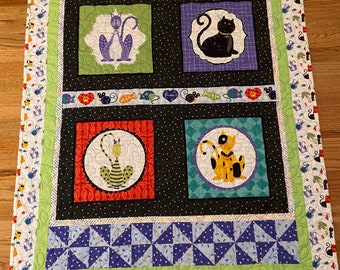 May include: A black and white quilt with a patchwork design featuring four squares with different colored backgrounds and images of cats. The squares are surrounded by a border of green, blue, and white fabric with a repeating pattern of cats and mice.